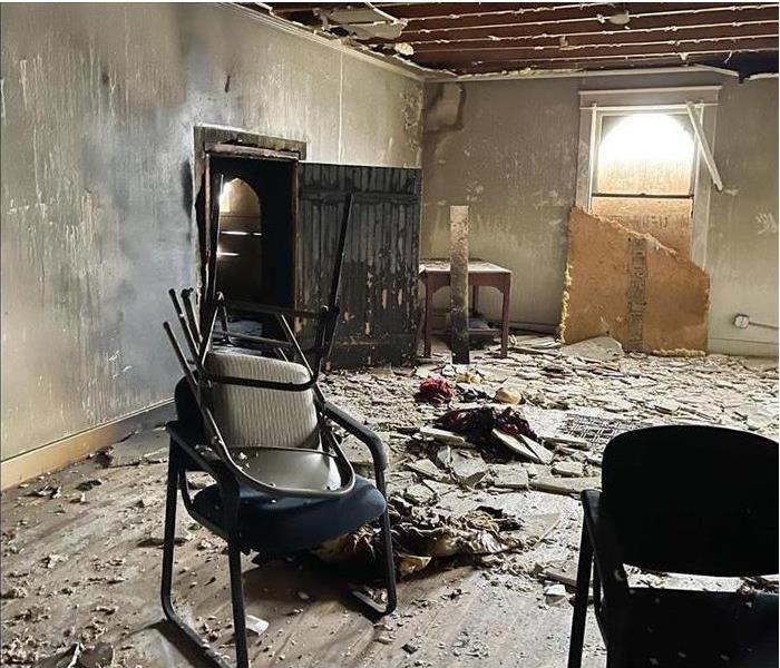 The walls inside a home are burned, ceiling has collapsed, debris on the floor, chairs